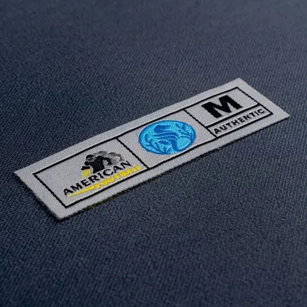 Rectangular woven label in grey and black reading American Authentic, size M, shown on navy fabric