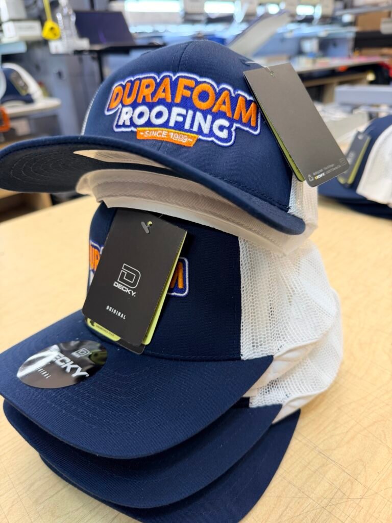 Navy and white trucker caps with raised puff embroidered roofing company logo, on the shop floor in Phoenix